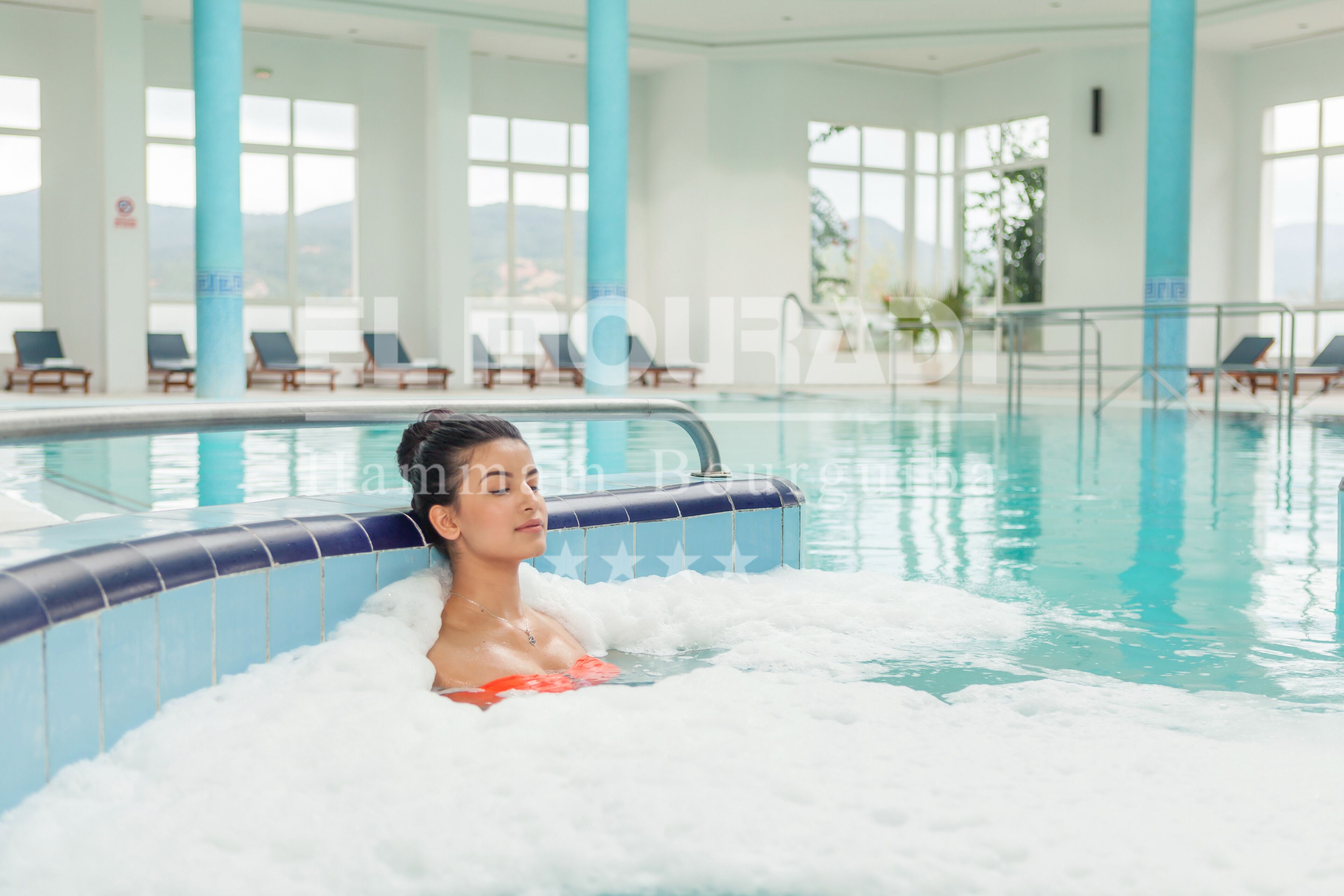Une personne se trouve dans un bain à remous, dans un grand espace de piscine intérieure aménagé avec des chaises longues et offrant une vue sur l’extérieur.