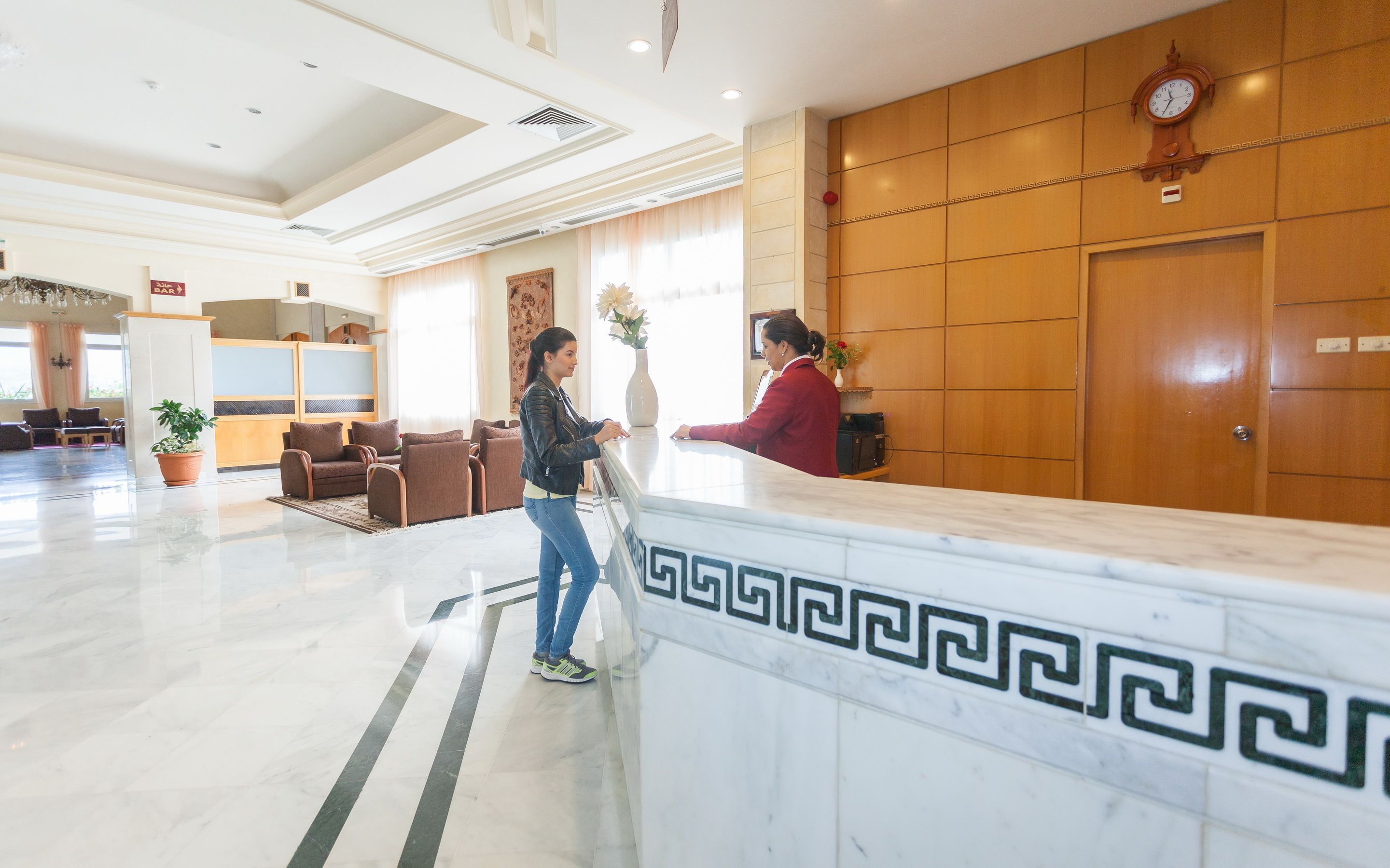 Un hall d’hôtel avec un comptoir de réception en marbre, un réceptionniste et un espace d’attente comprenant des canapés et une plante en pot.