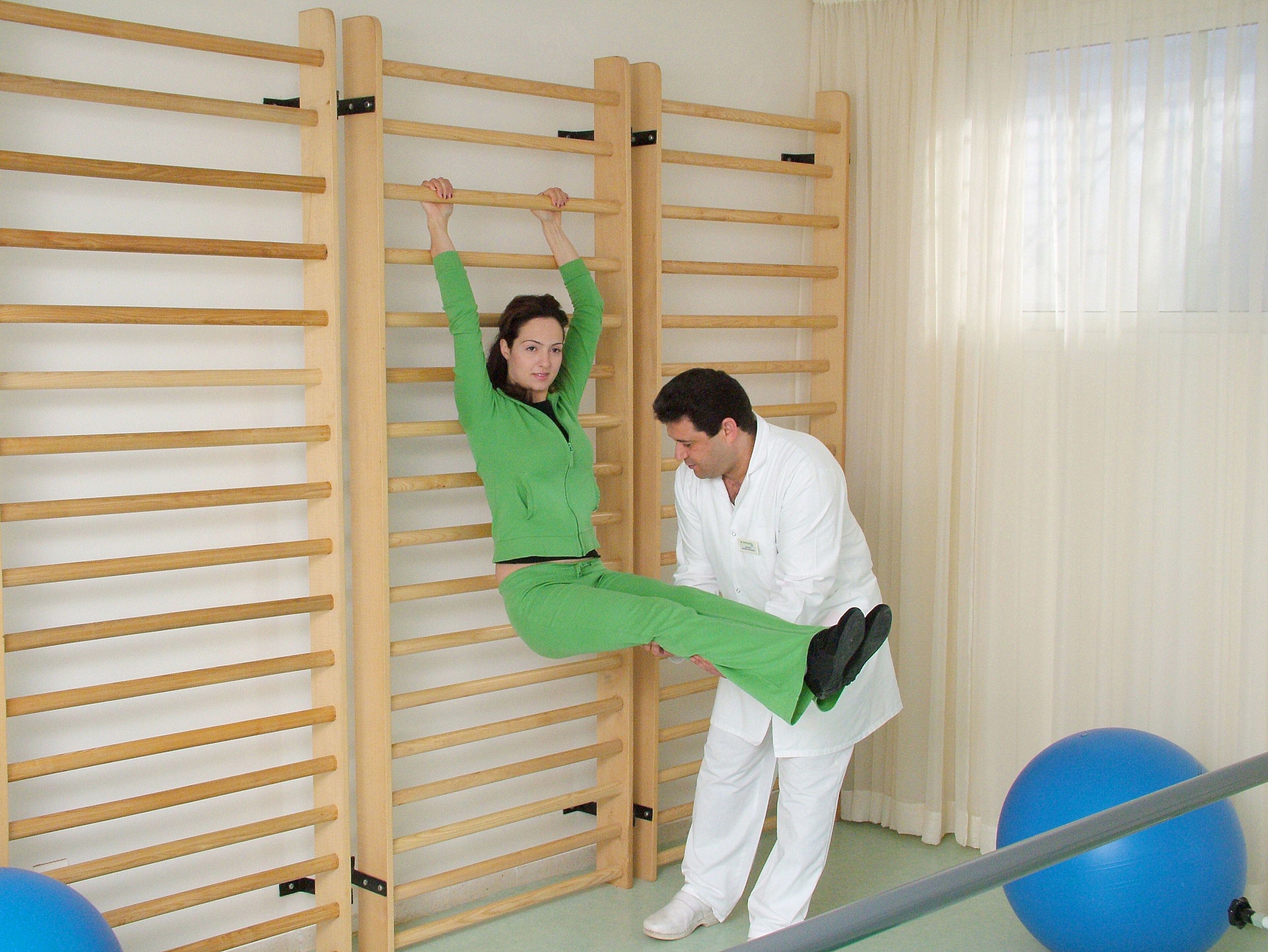 A person in green exercising on a wooden bar in a room with a large blue exercise ball and a window with curtains.