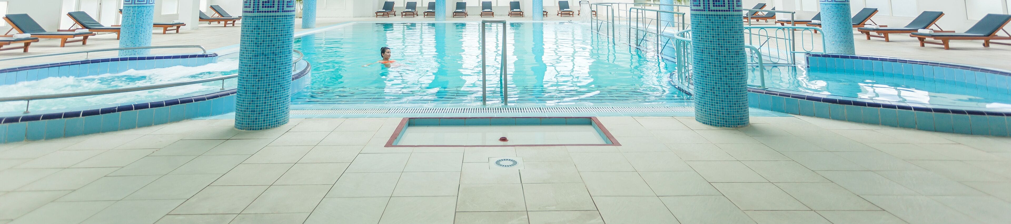 An indoor swimming pool with tiled walls and a central lane, surrounded by lounge chairs and a tiled floor.