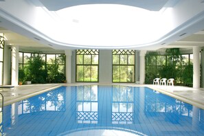 An indoor swimming pool with clear blue tiles, large windows with green foliage outside, and white chairs along the poolside. - El Mouradi Douz (Douz Nord)