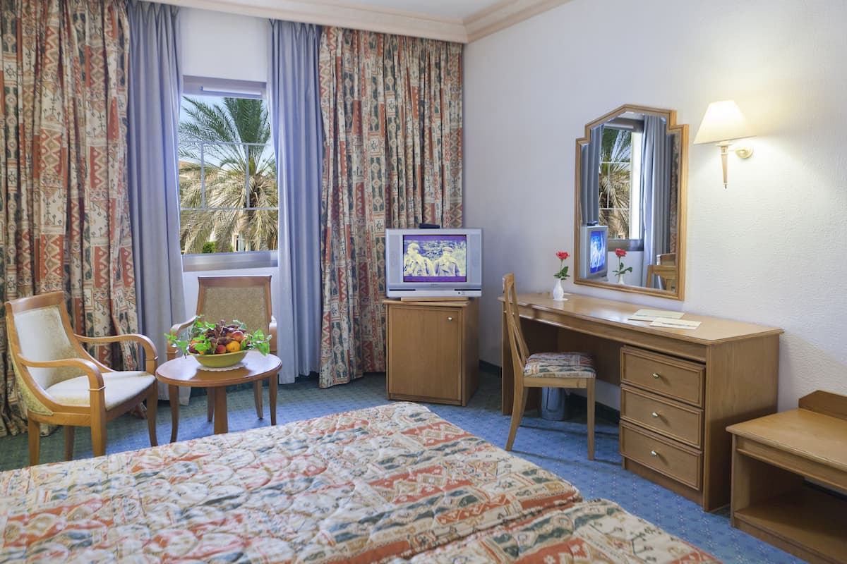A hotel room with a bed, a desk with a television, a chair, a small table with a fruit bowl, and a window with curtains.