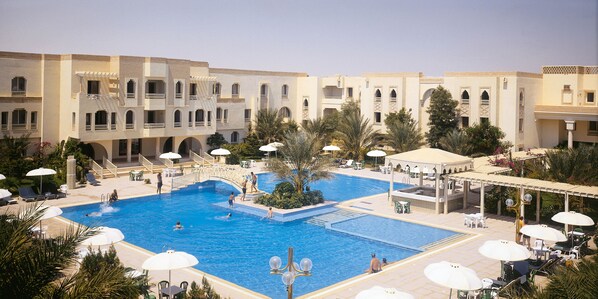A large outdoor swimming pool surrounded by lounge chairs and umbrellas, with a building featuring multiple balconies and arches in the background. - El Mouradi Douz (Douz Nord)