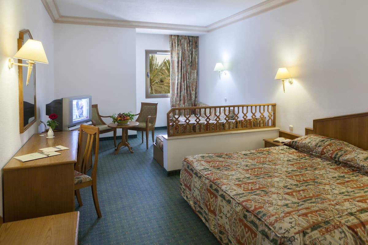 A hotel room with a bed, a desk with a computer, a chair, a small table with a bowl of fruit, and a window with curtains.