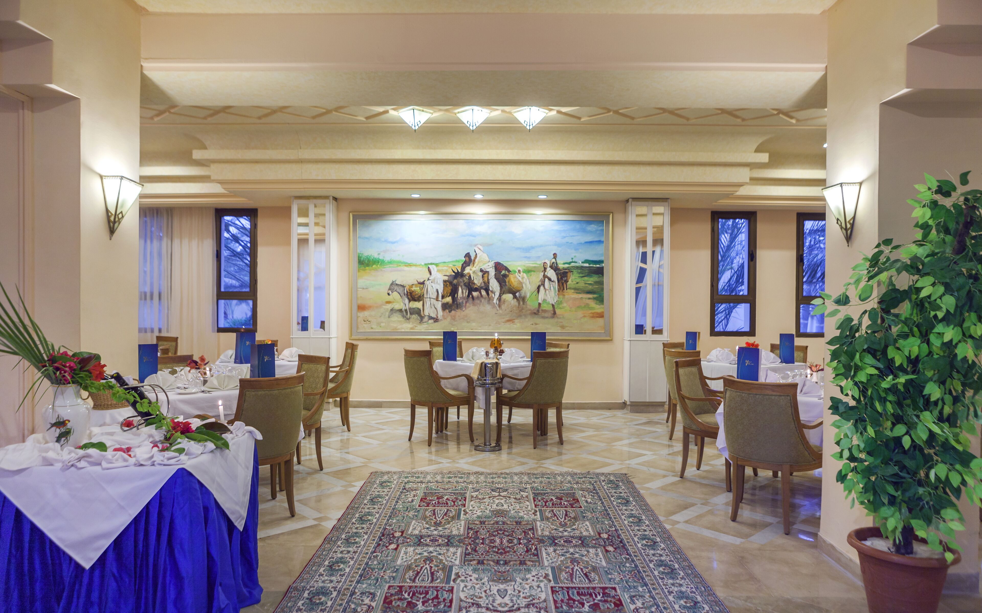 Un coin repas avec des tables dressées pour le repas, un grand tableau accroché au mur et un tapis à motifs sur le sol.