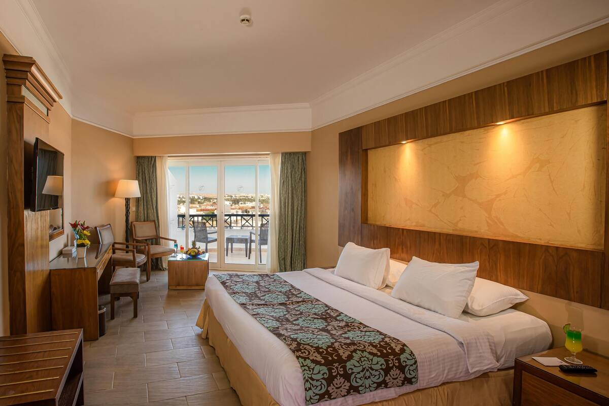 A hotel room with a bed, a desk, a chair, a TV, and a balcony with outdoor seating.