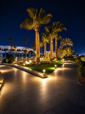 A nighttime view of a resort with palm trees, a paved walkway, and a building with multiple windows. - Naama Bay Hotel & Resort (Sharm El Sheikh)