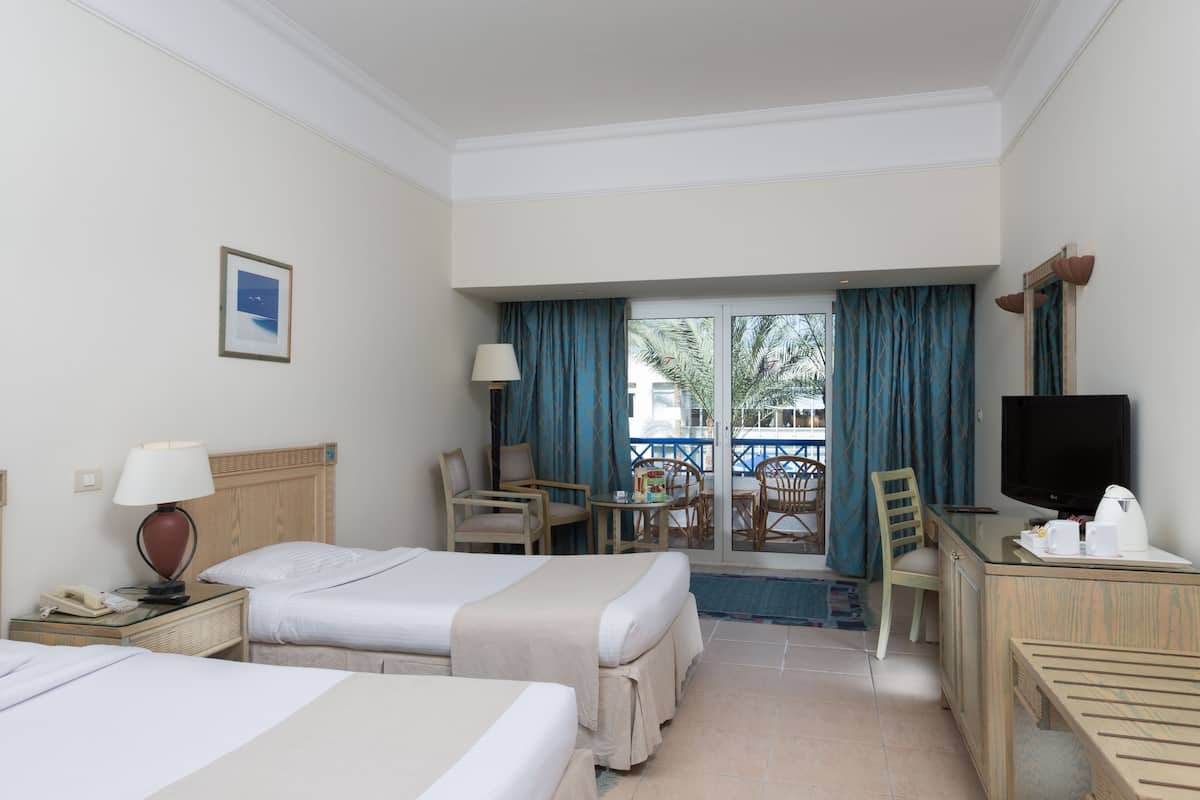 A hotel room with two beds, a desk with a TV, a phone, and a lamp. There is a balcony with a view of palm trees.