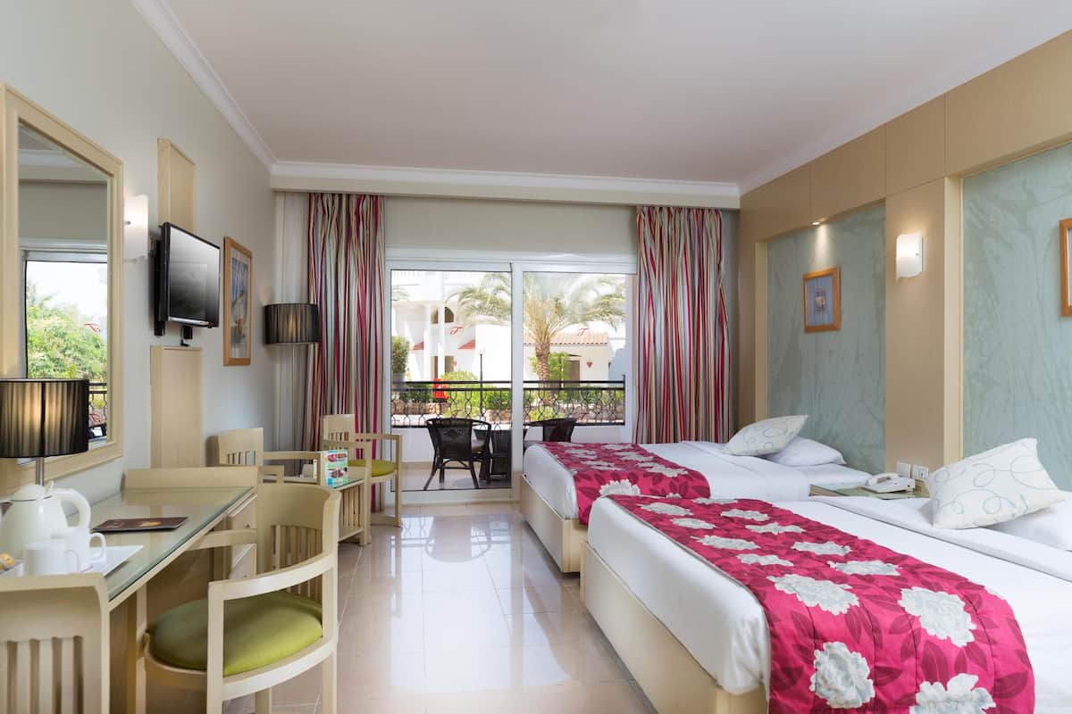 A hotel room with two beds, a desk with a chair, a TV, and a balcony with curtains.