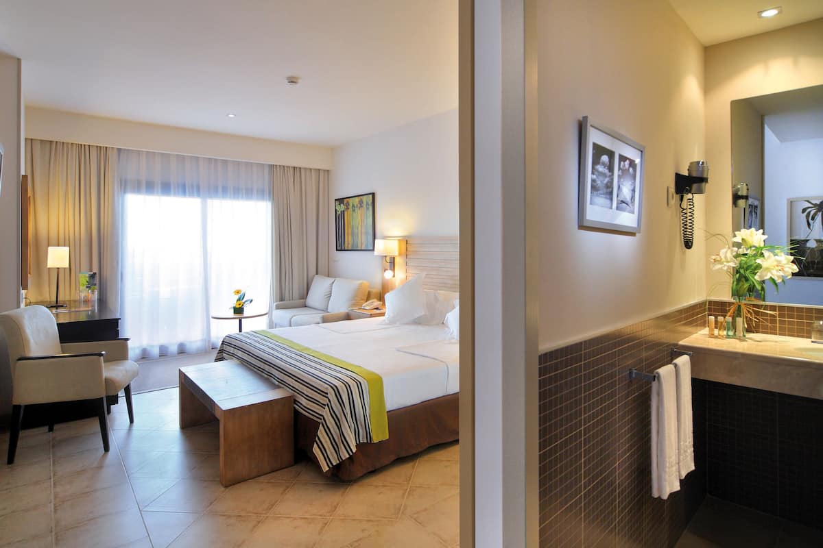 A hotel room with a bed, a desk, a chair, a small table with a lamp, a bathroom with a sink and mirror, and a picture on the wall.