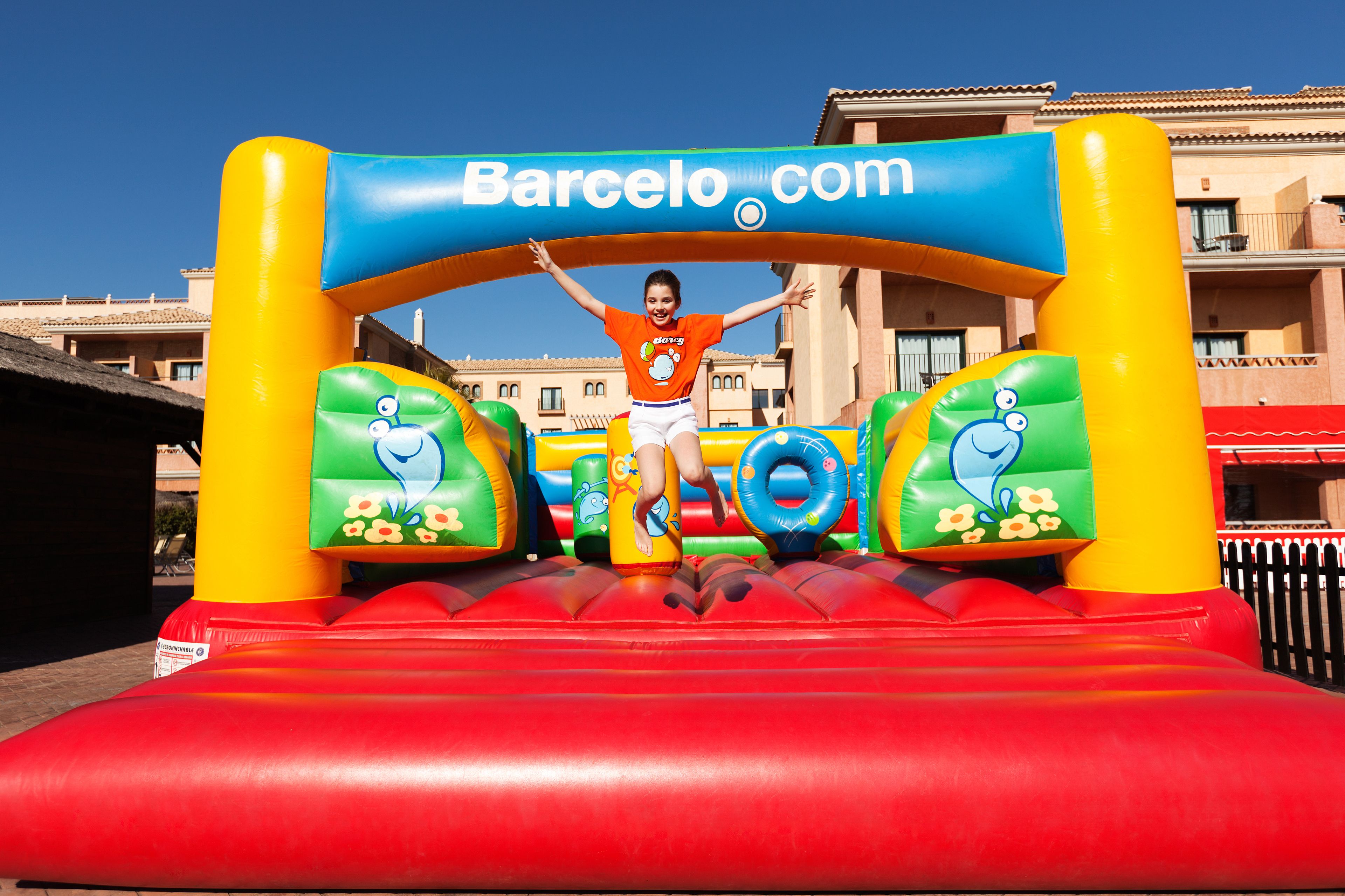 Värikäs, ilmatäytteinen rakennelma, jonka päällä lapsi hyppii. Rakennelmassa on logo ja teksti: ”Barcelo.com”.