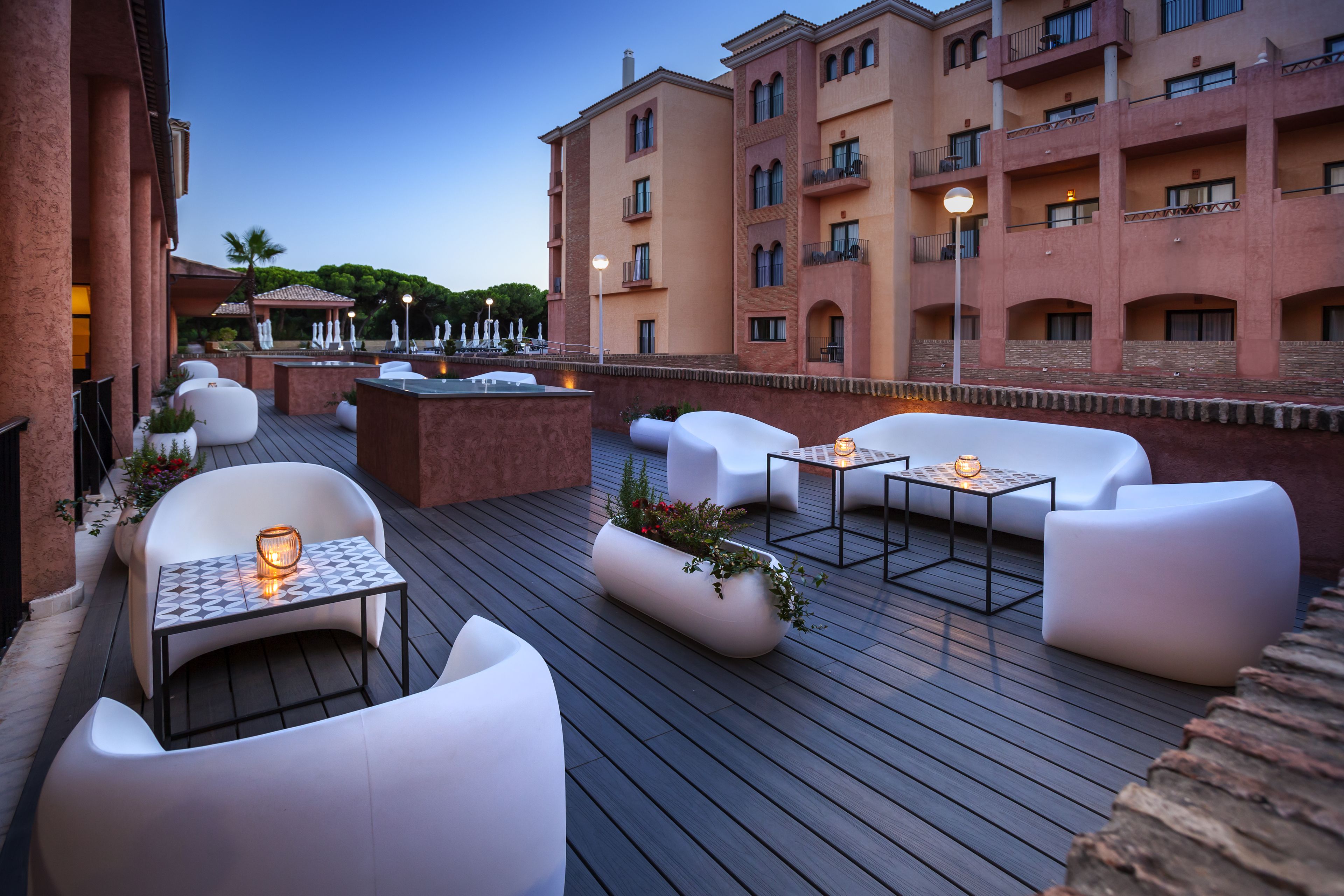 A rooftop patio with white outdoor furniture, a small table with a candle, and a planter with flowers.