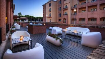 A rooftop patio with white outdoor furniture, a small table with a candle, and a planter with flowers.