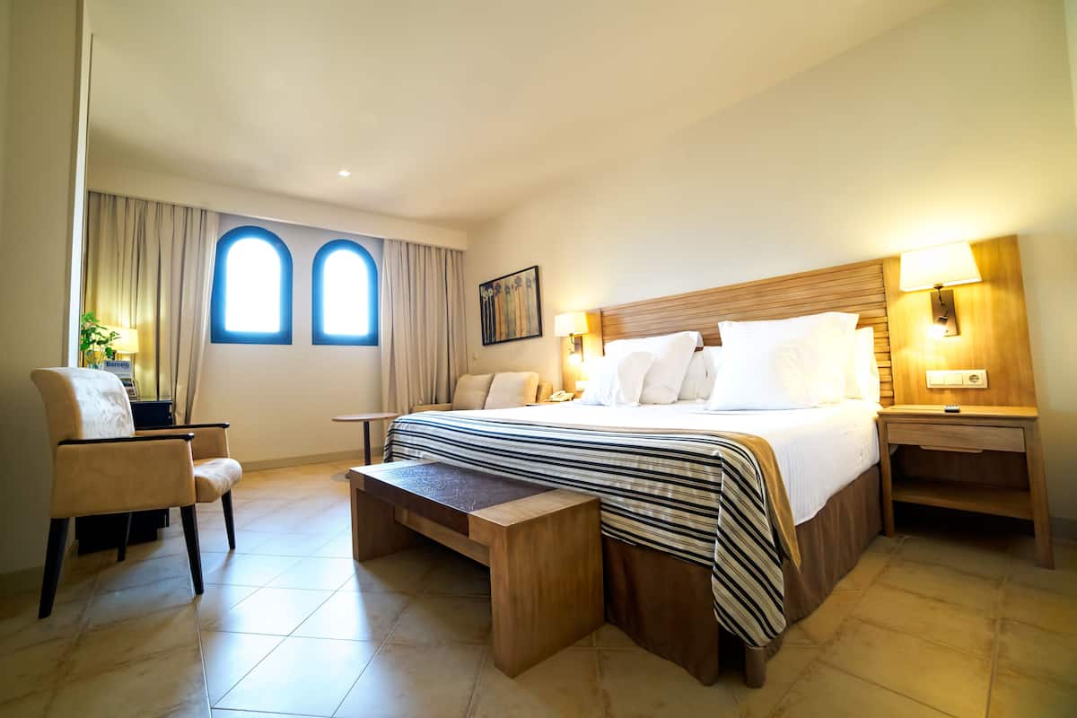 A hotel room with a large bed, two bedside lamps, a wooden nightstand, a bench, a chair, and two windows with curtains.