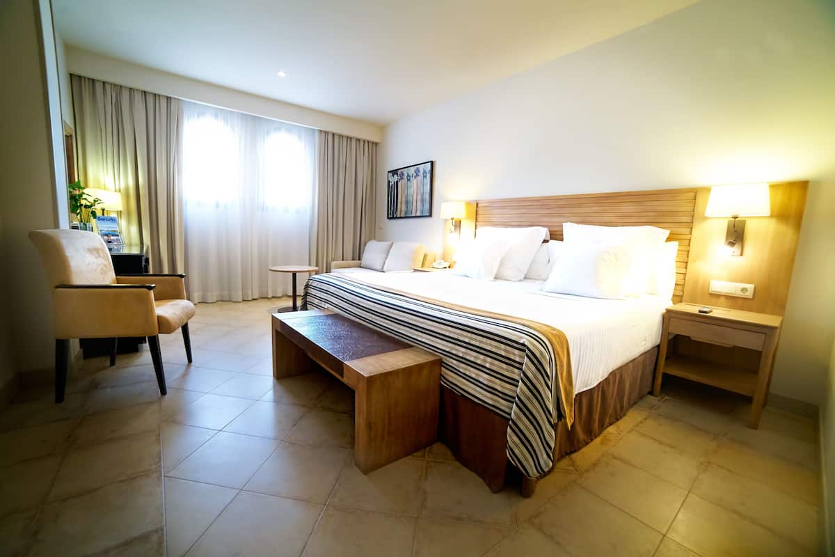 A hotel room with a large bed, a wooden bench, a desk, a chair, and a nightstand with a lamp.