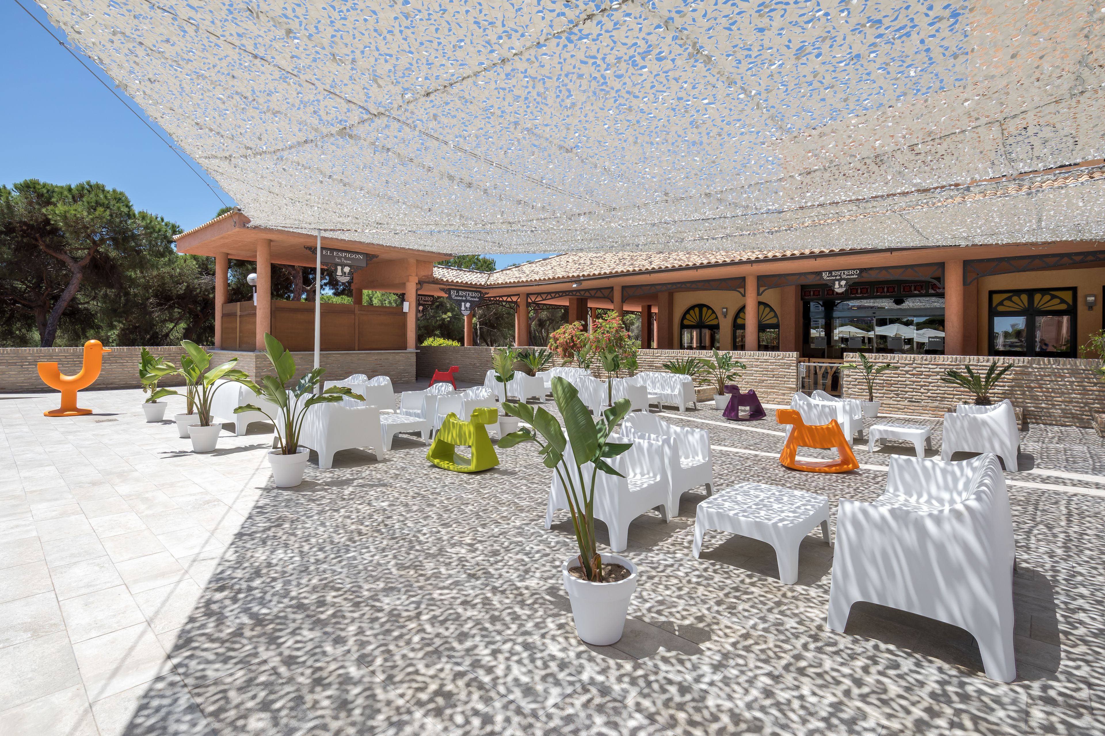Un área de estar al aire libre con sillas de plástico blanco y plantas en macetas, bajo un toldo decorativo.