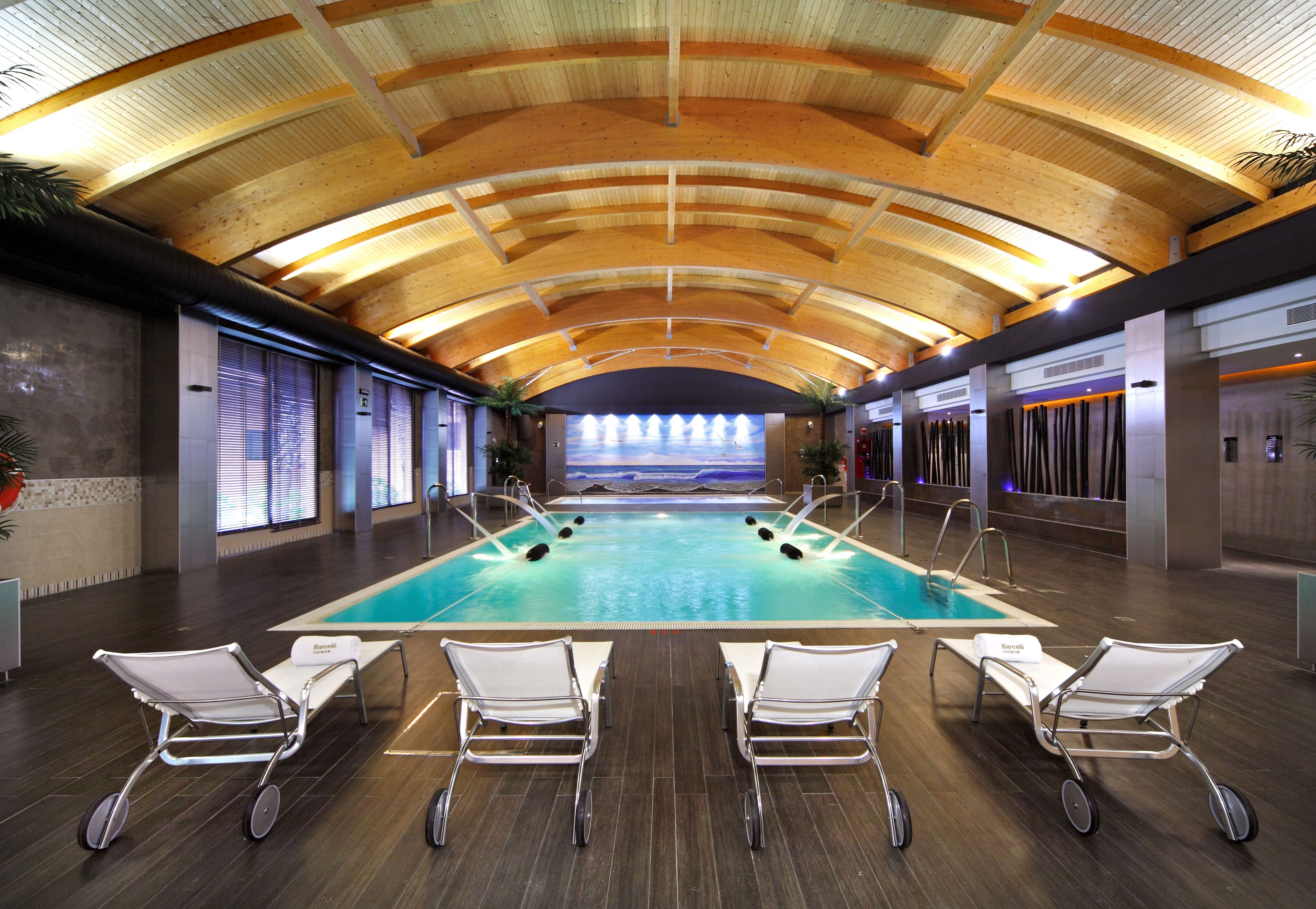Una amplia piscina interior con techo de madera, rodeada de sillones blancos.