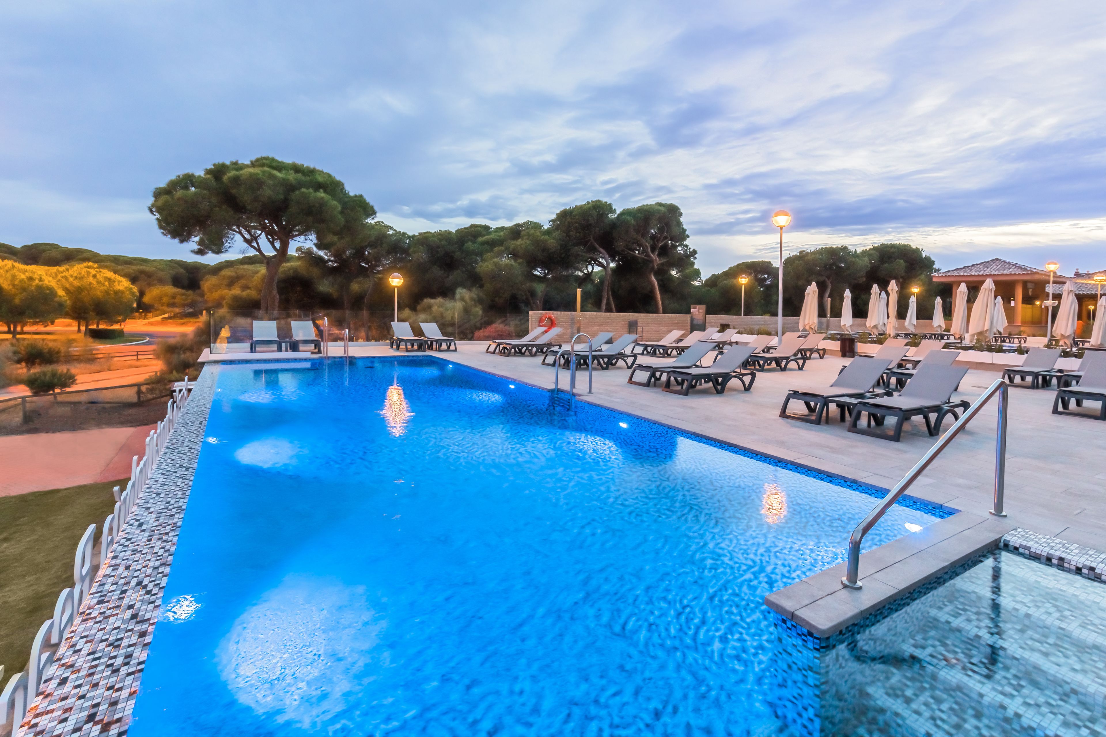 A swimming pool with lounge chairs and umbrellas, surrounded by trees and a building in the background.