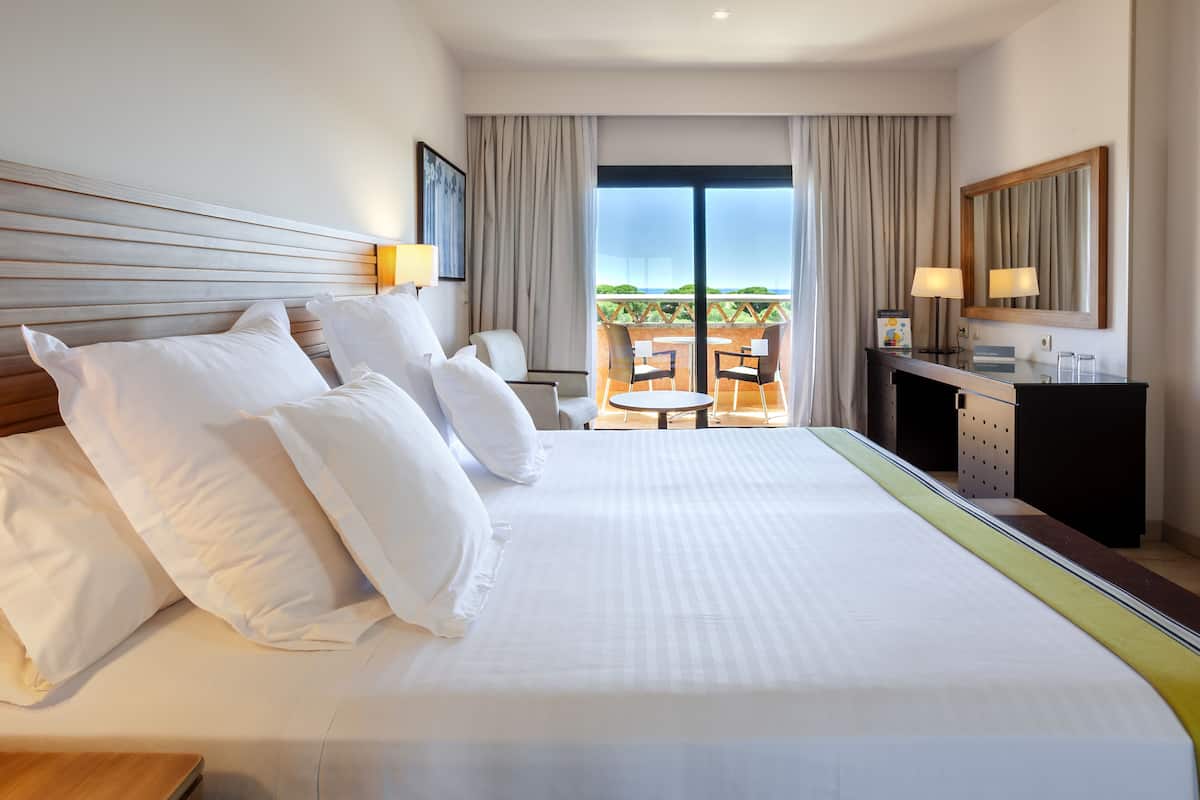 A hotel room with a large bed, a desk with a chair, a small table with chairs, and a balcony with a view.