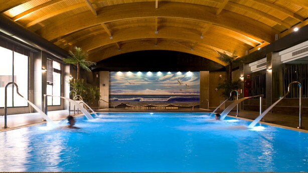 An indoor swimming pool with a mural of a beach scene, wooden ceiling beams, and two water jets. - Barceló Punta Umbria Beach Resort (Punta Umbría)