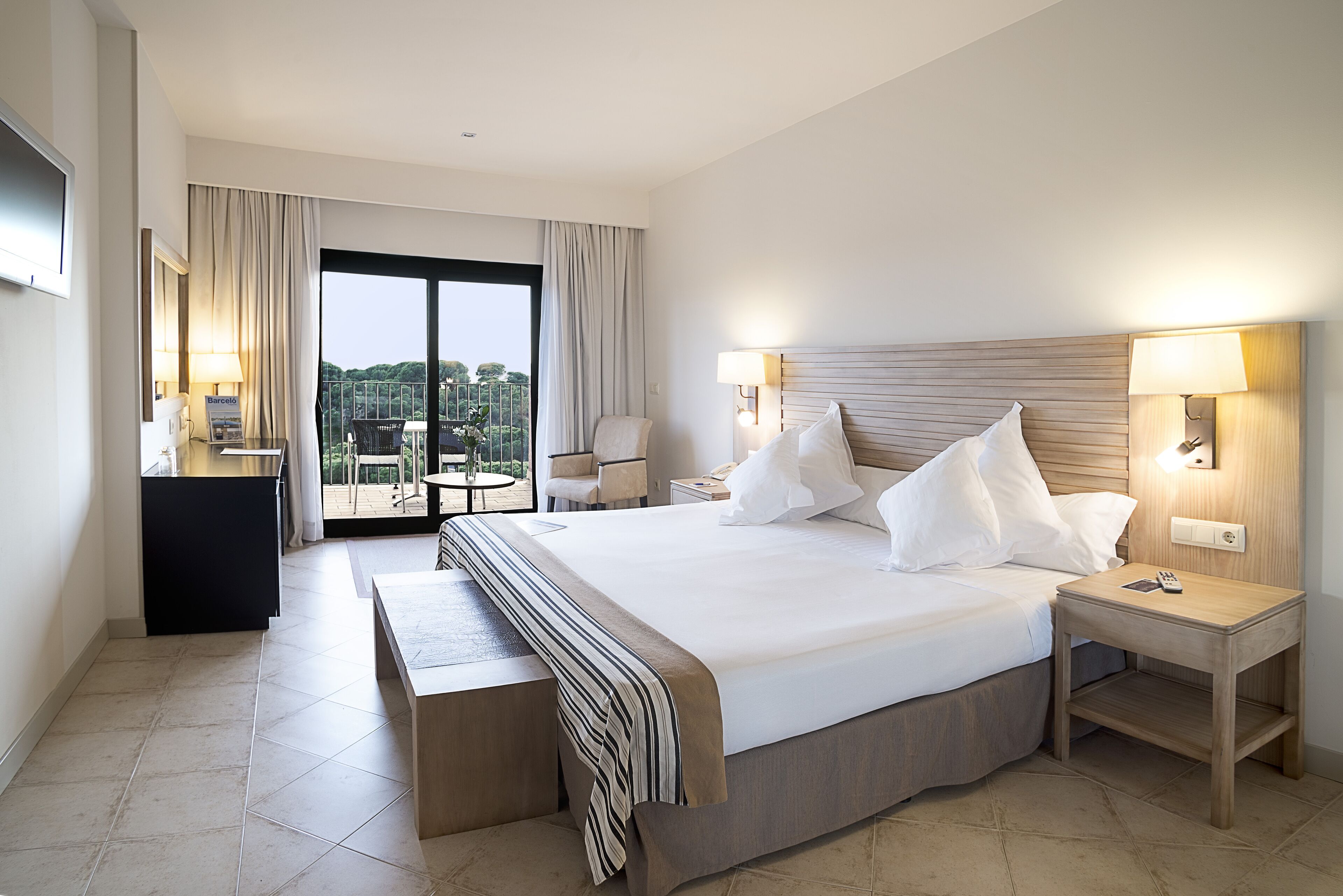 A hotel room with a large bed, bedside tables, a bench, a desk with a lamp, and a view of the outdoors.