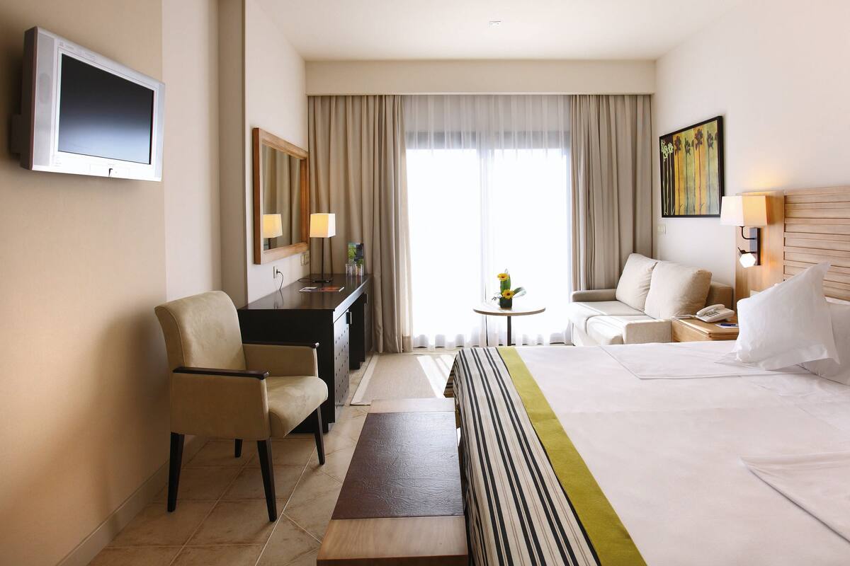 A hotel room with a bed, two armchairs, a desk, a TV, and a painting on the wall.
