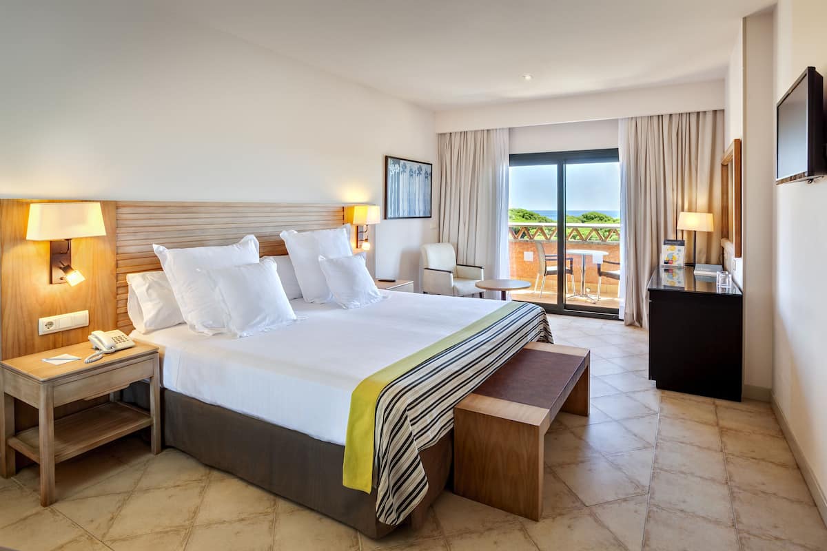 A hotel room with a large bed, bedside tables, a desk, a chair, a TV, and a balcony with a view.