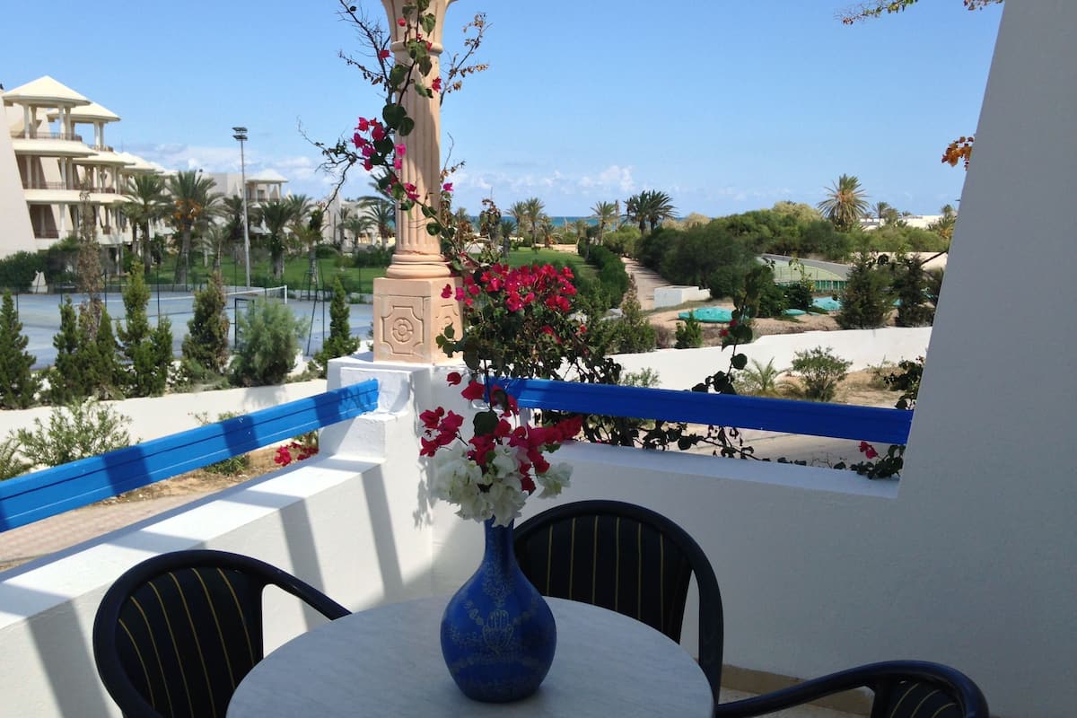 En balkon med et bord, stole, en vase med blomster og udsigt til en pool og bygninger.