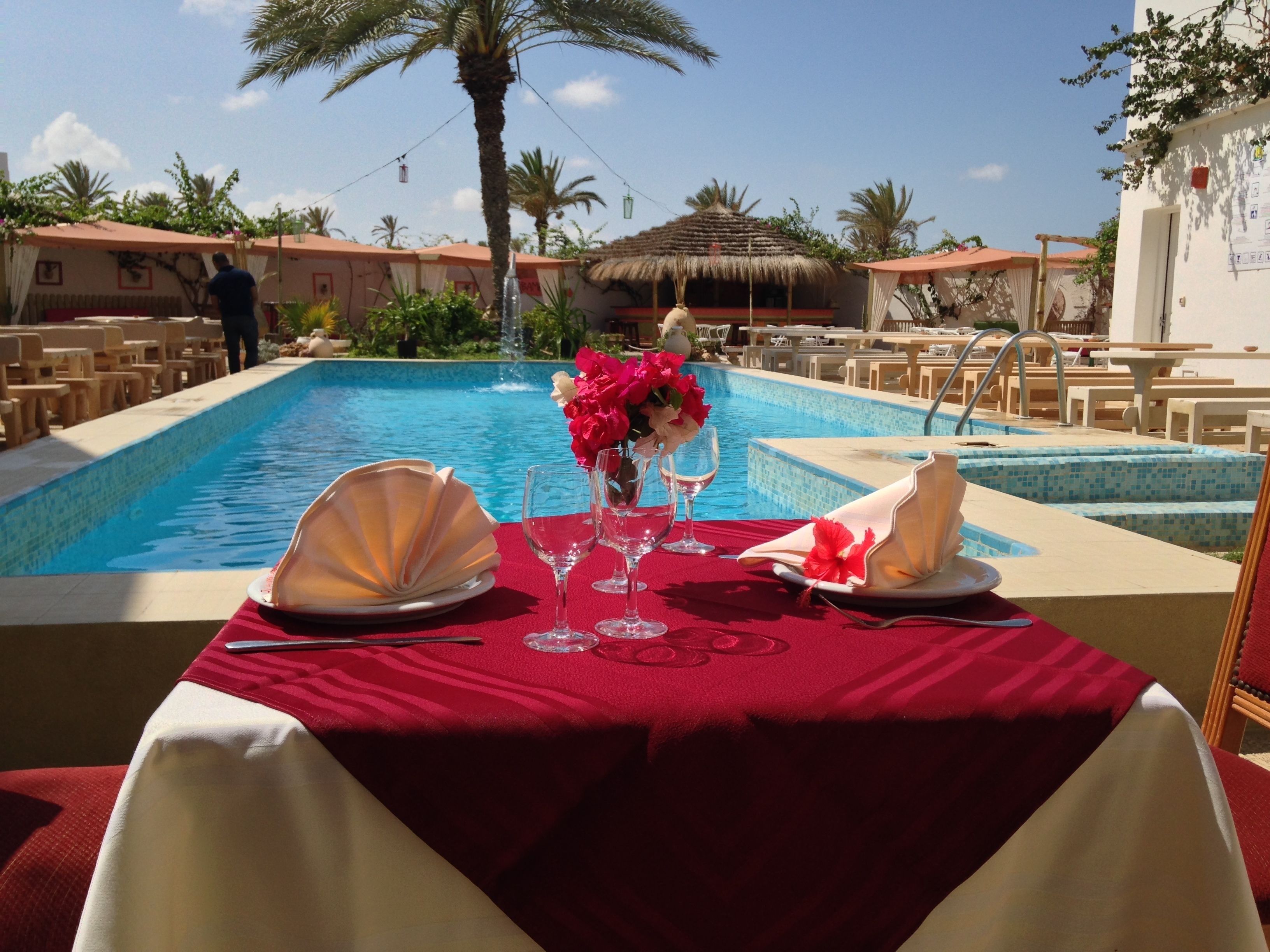 A dining table set for two with a red tablecloth, two white plates, two wine glasses, and a vase of flowers.