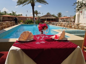 A dining table set for two with a red tablecloth, two white plates, two wine glasses, and a vase of flowers.