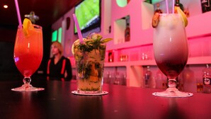 Three cocktails on a bar counter with a TV in the background. - Rodes (Djerba Midun)
