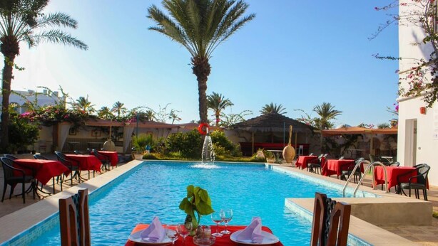 A poolside dining area with tables set for two, surrounded by palm trees and a clear blue pool. - Rodes (Djerba Midun)