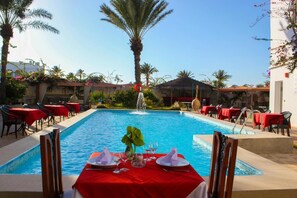 A poolside dining area with tables set for two, surrounded by palm trees and a clear blue pool.