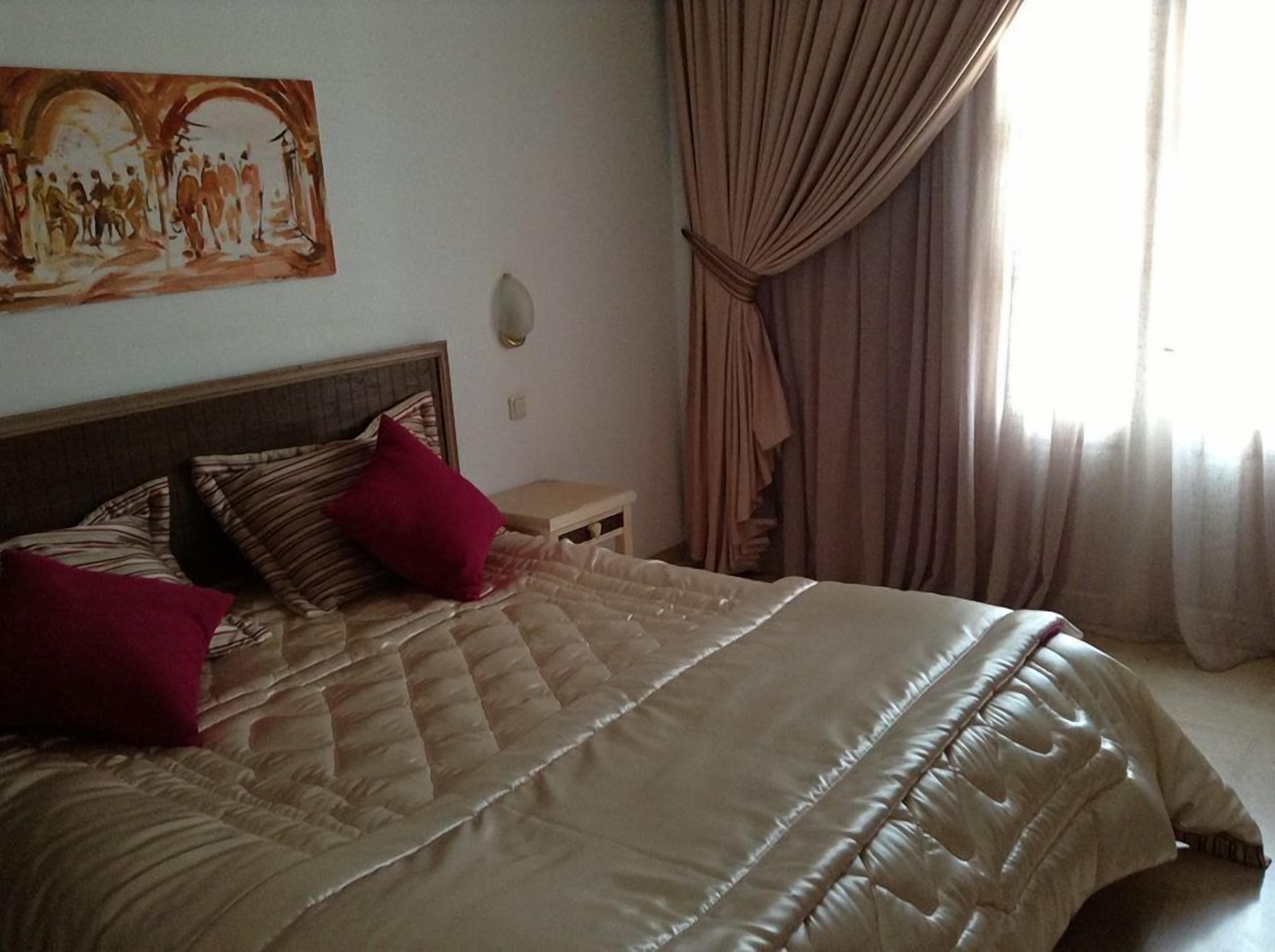 A bed with a quilted cover, two red pillows, a wooden headboard, a small bedside table, and a painting on the wall.