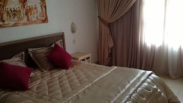 A bed with a quilted cover, two red pillows, a wooden headboard, a small bedside table, and a painting on the wall.