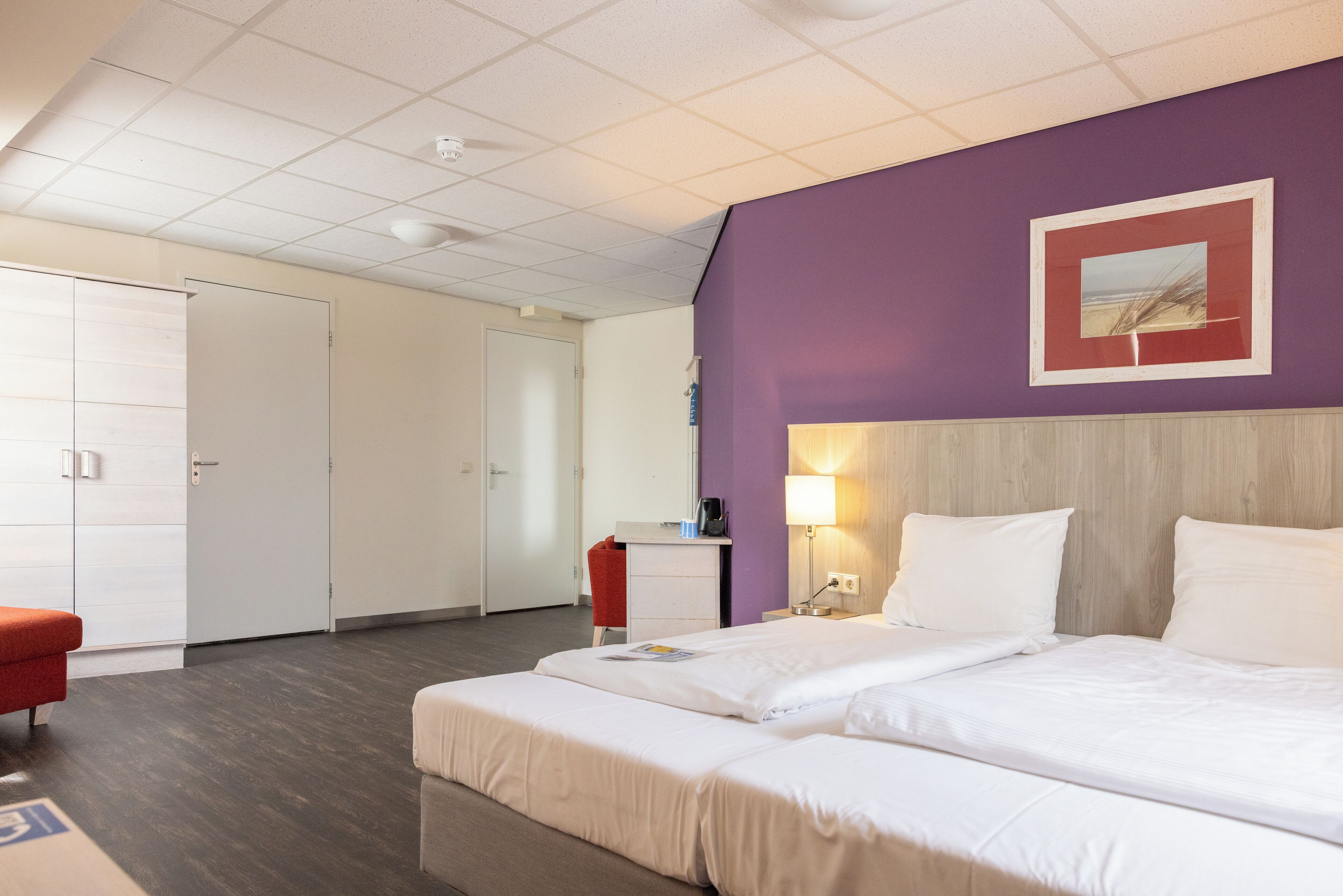 A hotel room with a large bed, a desk, a chair, and a purple accent wall.