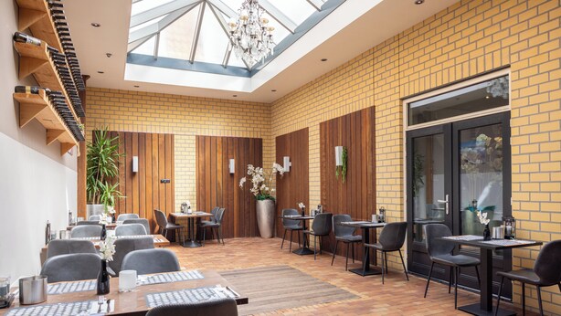 Un vaste espace de restauration intérieur, avec des murs en briques, des lambris en bois et une verrière. Les tables sont disposées avec des chaises, et une grande plante en pot est visible.