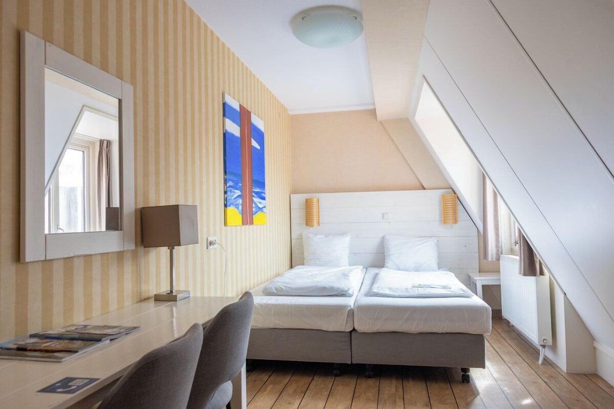 Ein Hotelzimmer mit Bett, Schreibtisch, Stuhl, Lampe, Spiegel und Fenster.