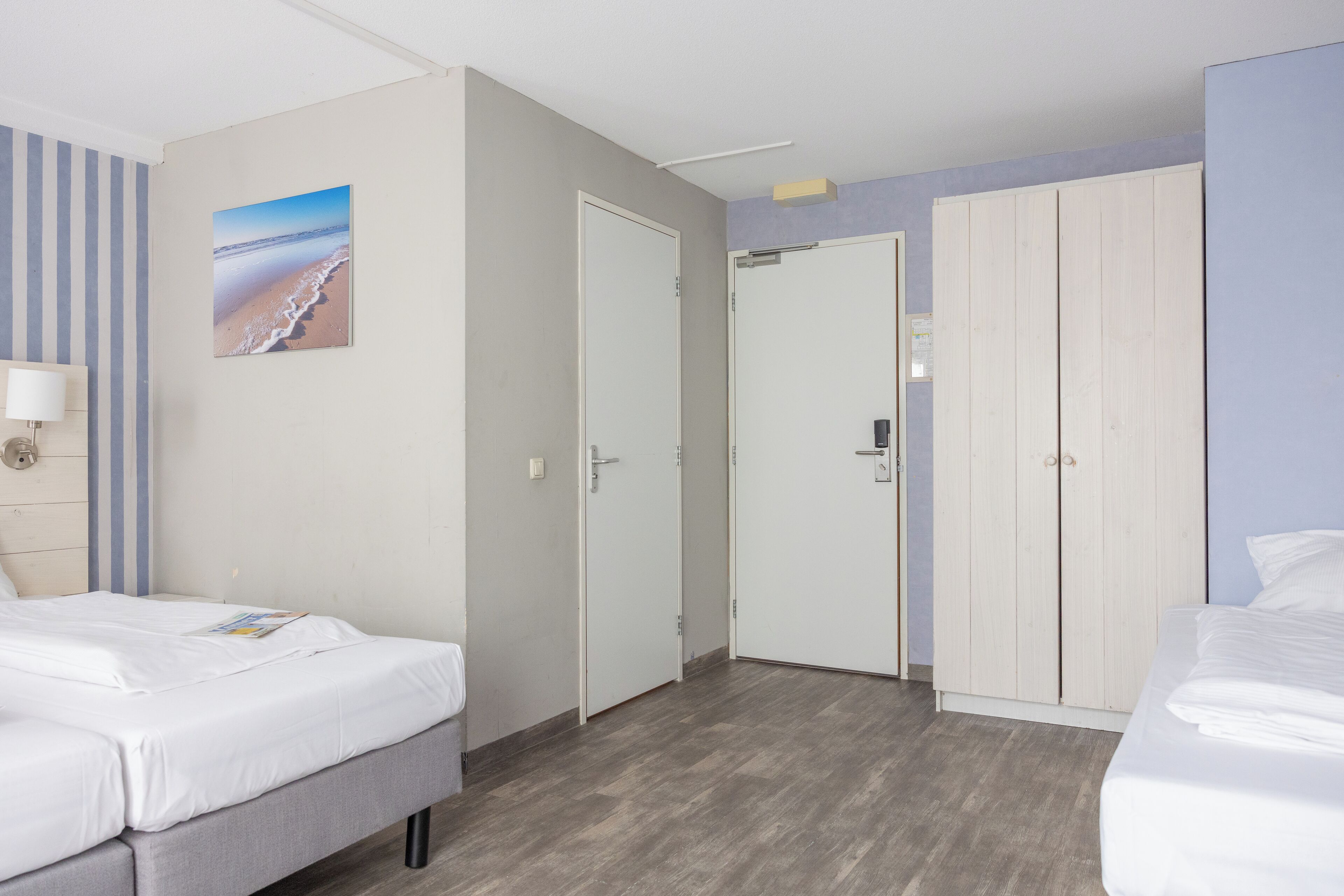 A hotel room with two beds, a large wardrobe, a painting of a beach, and a striped wall.