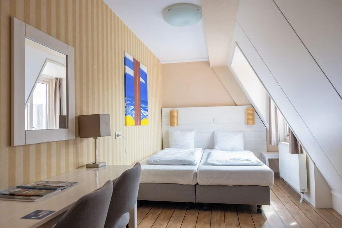 Une chambre d’hôtel avec un lit, un bureau, une chaise, une lampe, un miroir et une fenêtre.