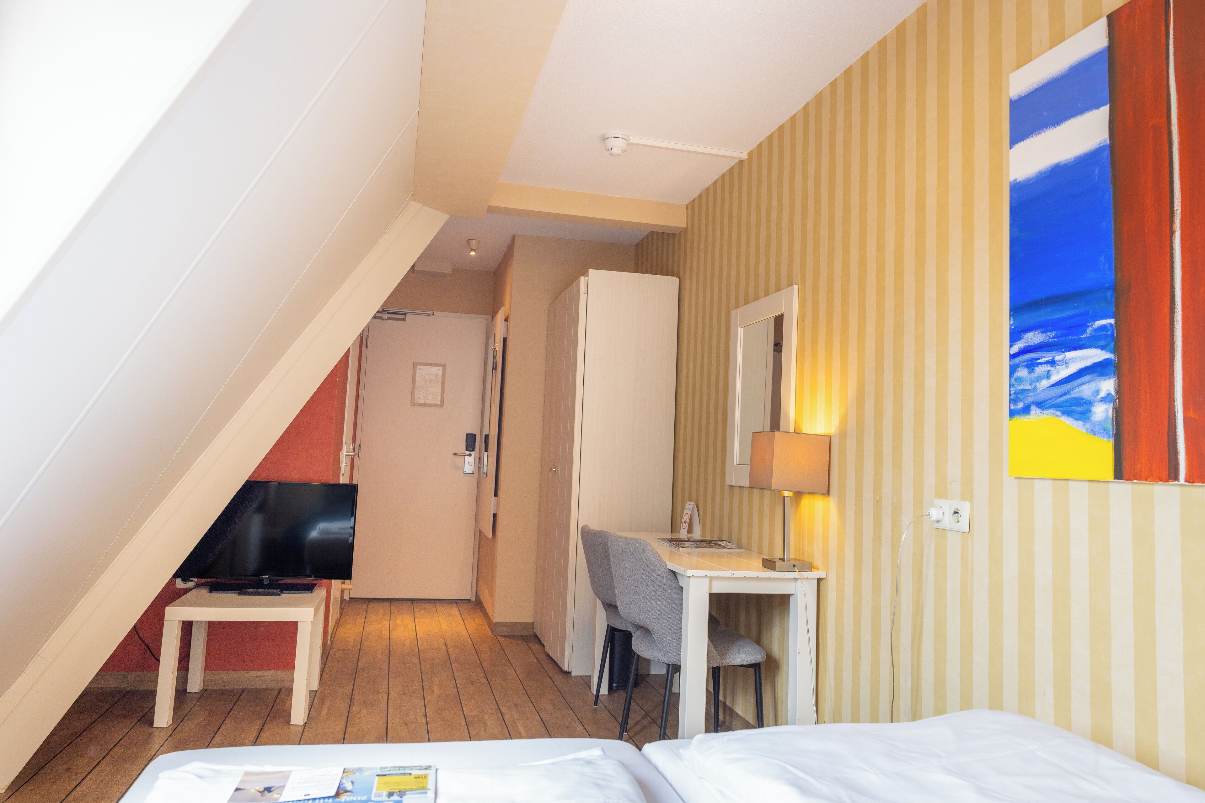 A hotel room with a bed, a desk, a television, and a painting of a beach scene.