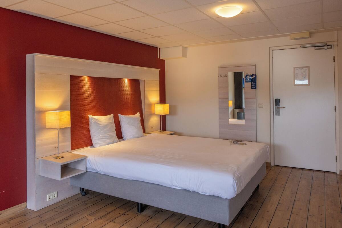 Une chambre d’hôtel avec un grand lit, deux tables de chevet avec des lampes, un plancher en bois et une porte dotée d’un miroir et d’un tableau d’affichage.