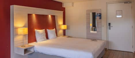 A hotel room with a large bed, two bedside tables with lamps, a wooden floor, and a door with a mirror and a notice board.