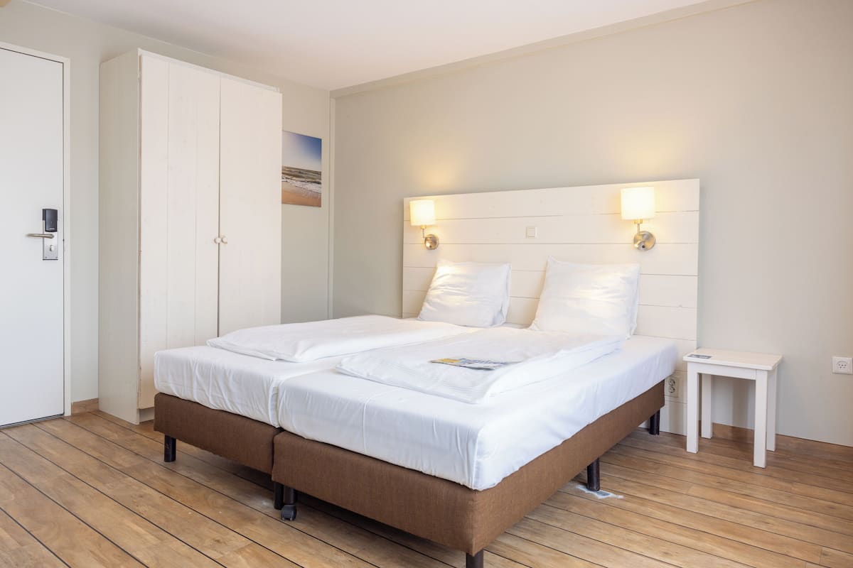 Un lit double avec une literie blanche, un plancher en bois, une armoire blanche et une table de chevet avec une lampe.