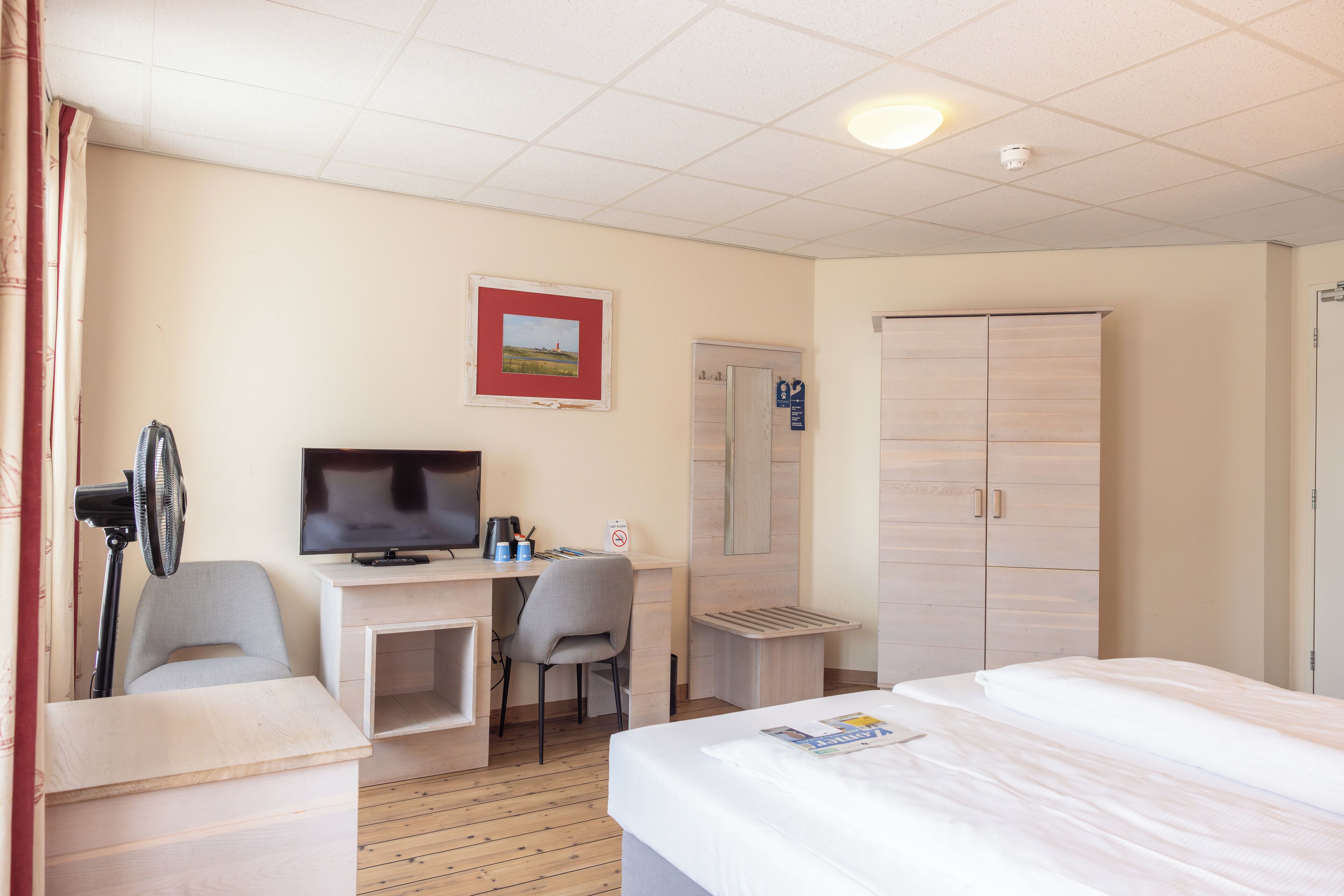A hotel room with a bed, a desk with a computer and a chair, a TV, a wardrobe, and a radiator.