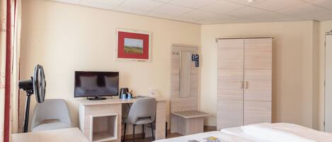A hotel room with a bed, a desk with a computer and a chair, a TV, a wardrobe, and a radiator.