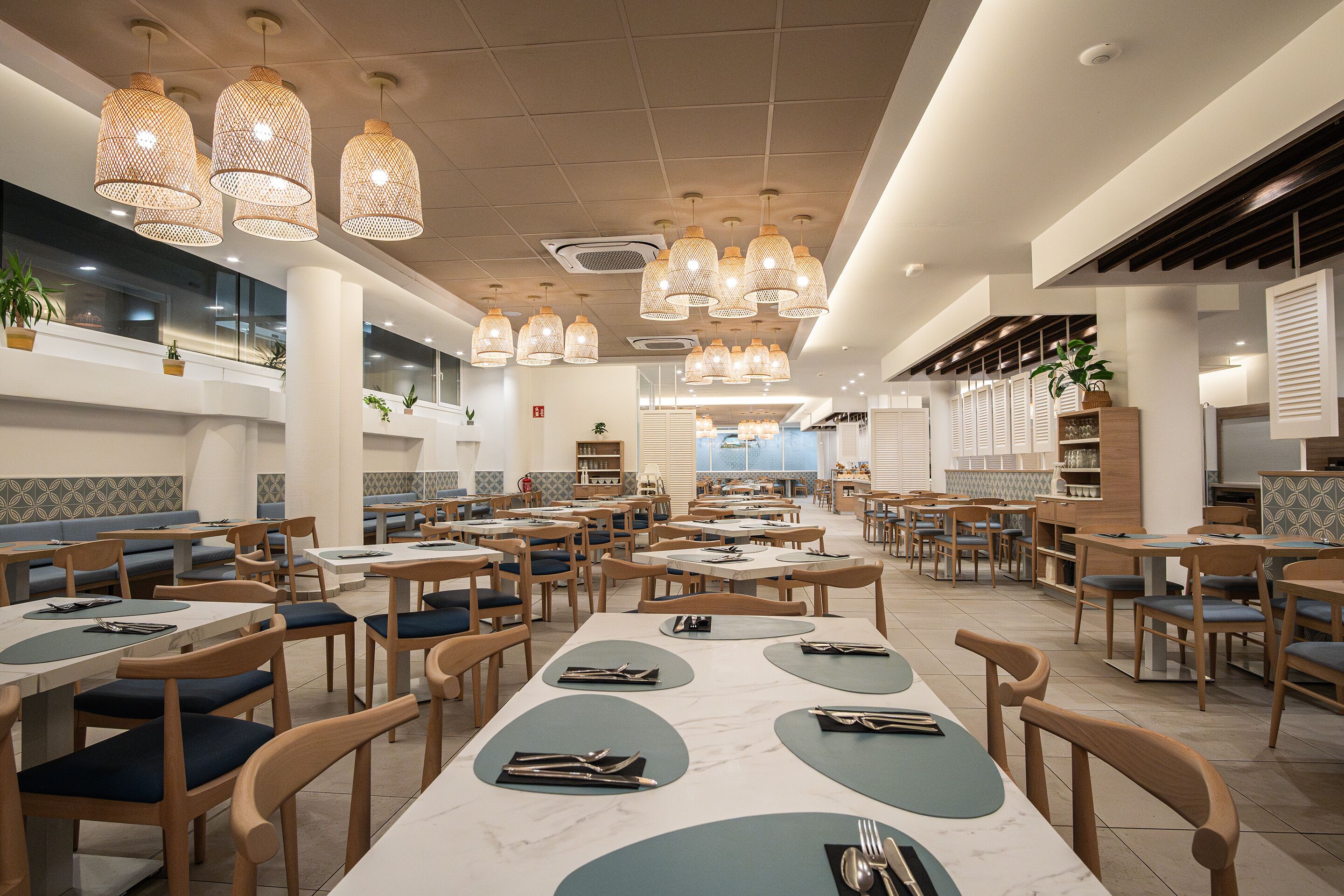 A spacious restaurant with multiple dining tables and chairs, overhead wicker pendant lights, and a modern interior design.