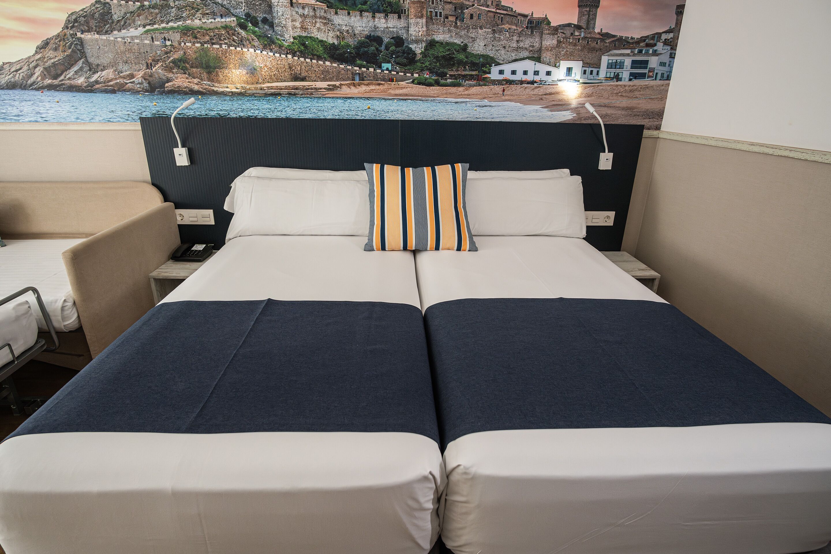 Cama doble con almohada a rayas, mesita de noche, una silla y vistas a un paisaje costero con un edificio histórico.