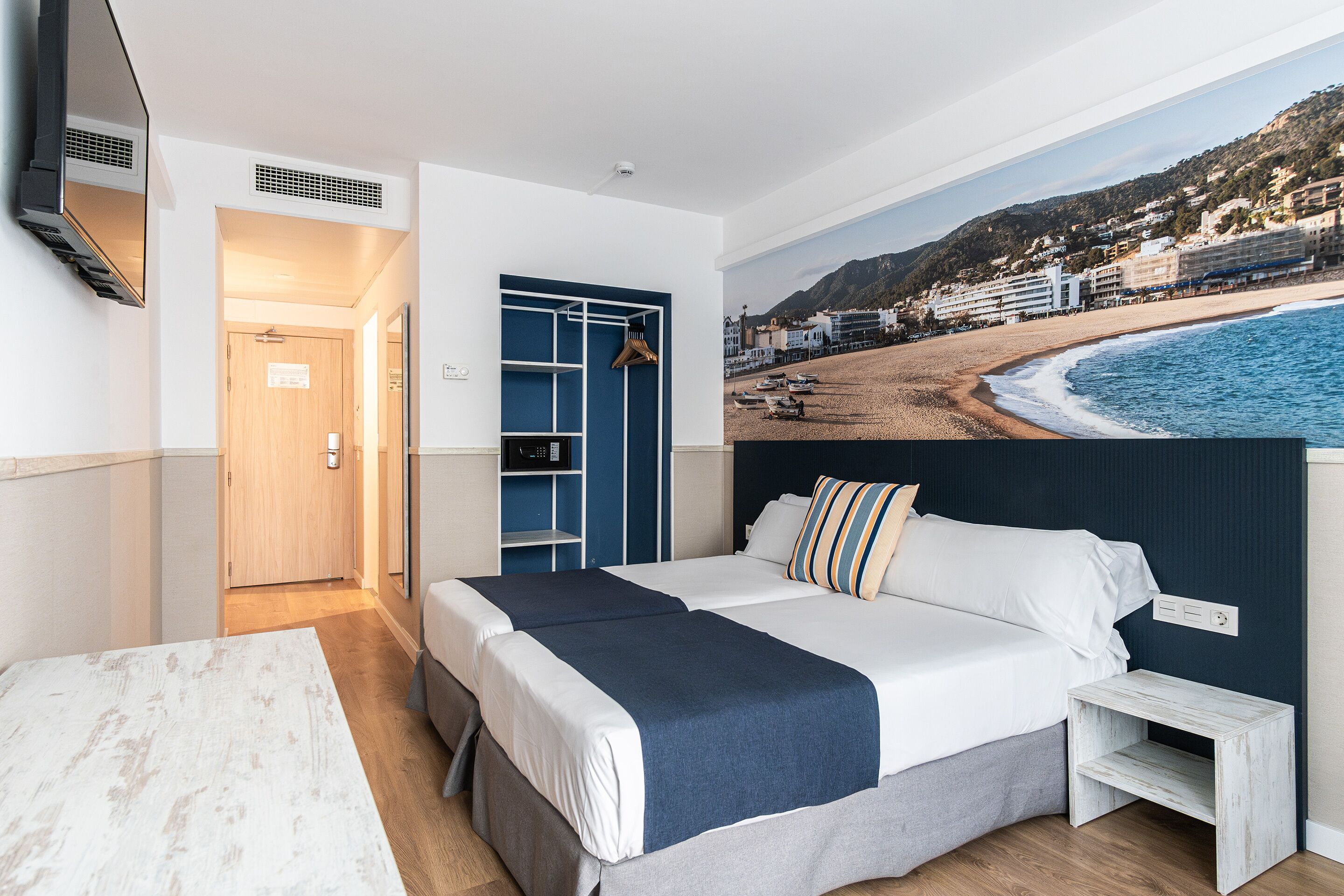 A hotel room with a bed, a desk, a wardrobe, and a large beach view poster.