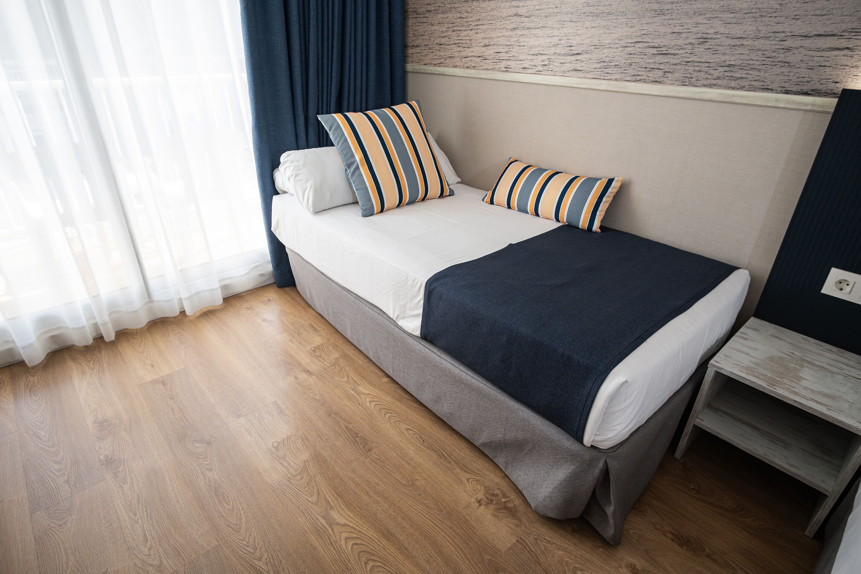 A neatly made bed with striped pillows, a dark blue blanket, and a wooden floor.